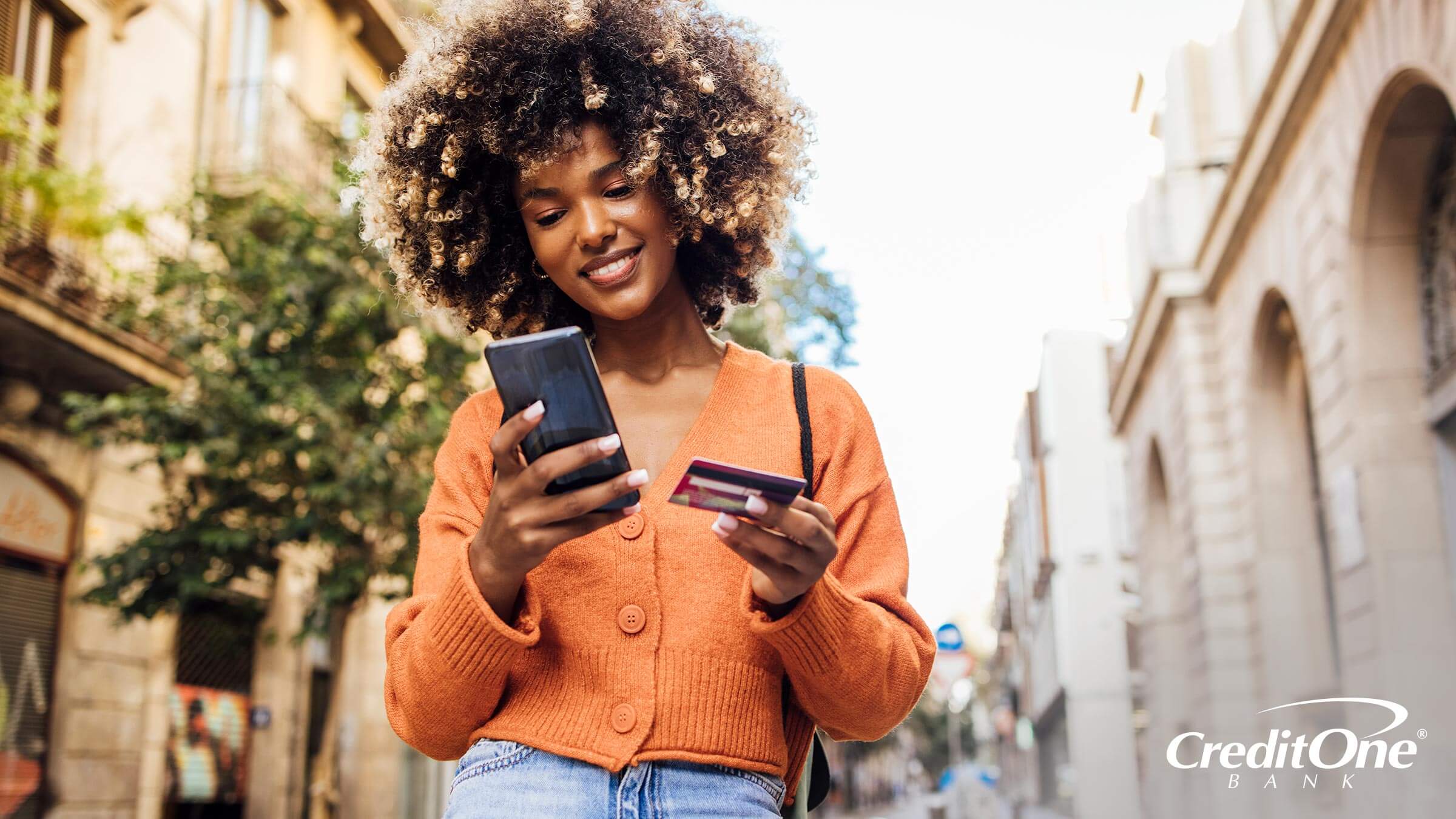 A young Gen Z woman walking down a quaint city street smiles as she uses her credit card to make a payment on her mobile phone.