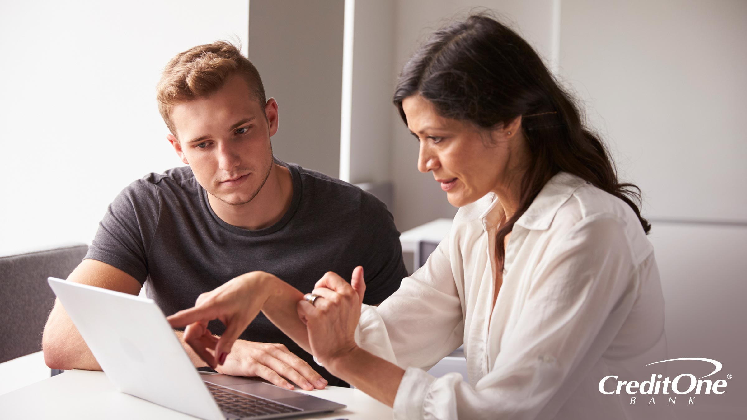 A young adult looks at his laptop while his mother points at the screen as if she’s walking him through the process of adding her as a cosigner.