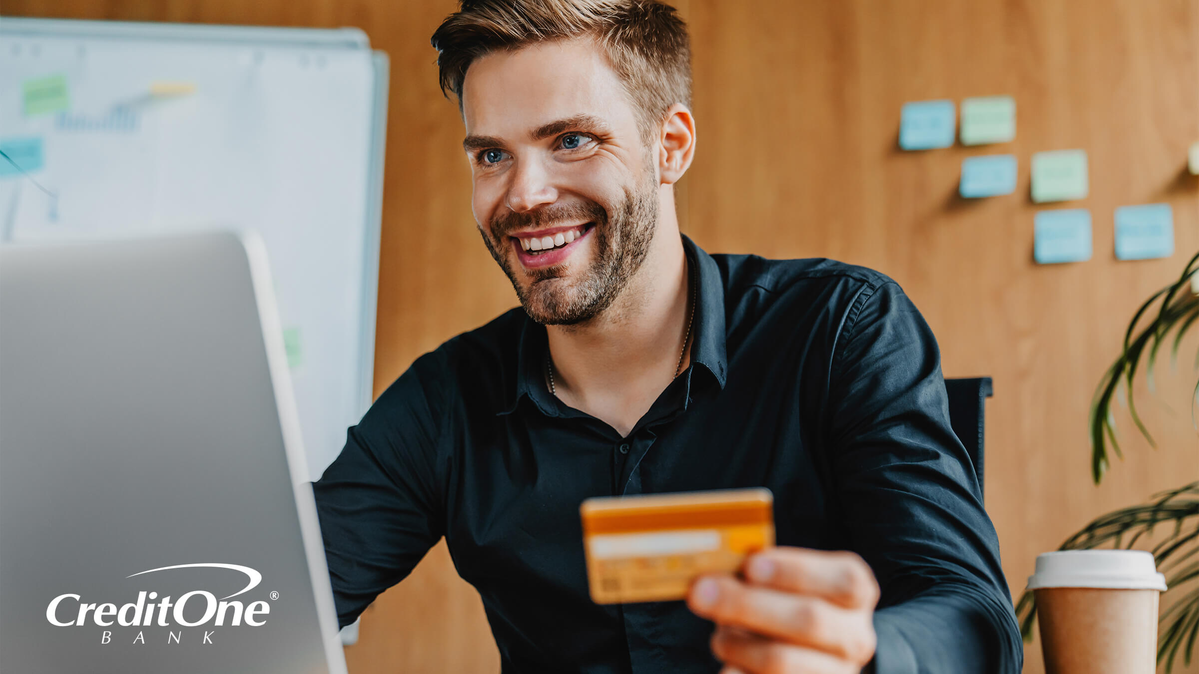 A young man smiles as he sits at his desk holding a credit card and using a laptop, which is a great tool for experiencing the benefits of online banking.