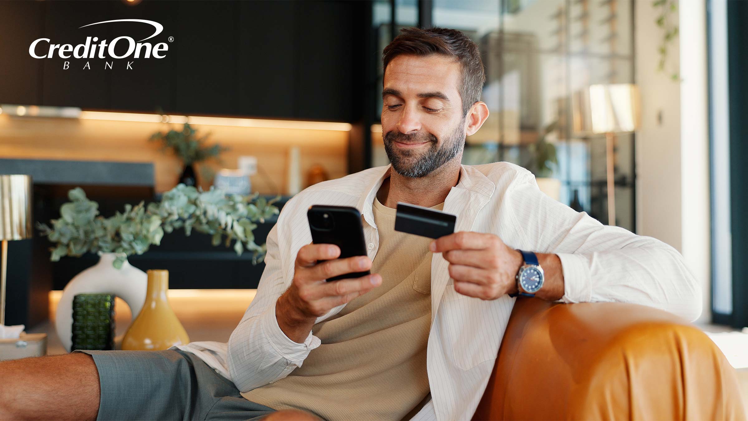 A man smiles while holding his credit card in one hand and his phone in the other. He may have received a notification about his secured credit card deposit, which is often refunded when the card becomes unsecured.