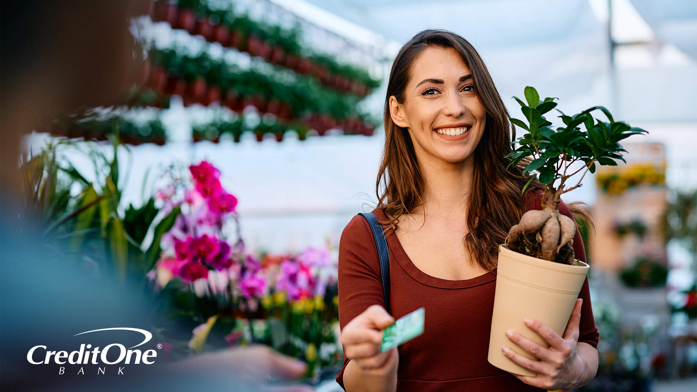 A smiling woman holds a potted plant in a retailer’s garden section, while presenting her store credit card for payment.